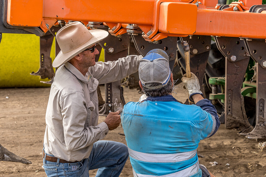 Two men are doing maintenance on a harvester. They are kneeling in the dirt tightening bolts.