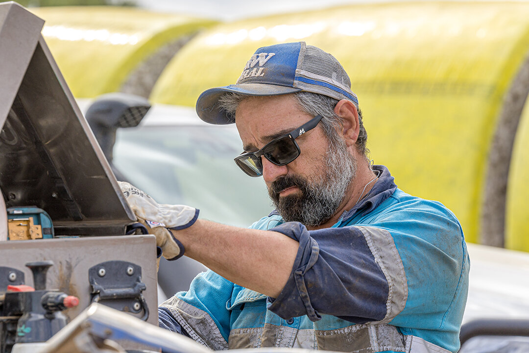 A man is doing maintenance on a machine. He is wearing a blue safety jacket and gloves